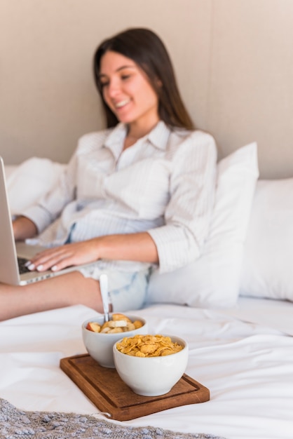 Souriante jeune femme à l&#39;aide d&#39;un ordinateur portable avec cornflakes et salade de fruits au lit