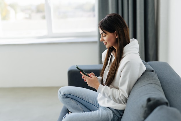 Souriante Jeune Femme à L'aide De Téléphone Assis Sur Un Canapé à La Maison En Regardant L'écran Du Smartphone Belle Femme Discutant Sur Les Réseaux Sociaux Ou Faisant Des Achats En Ligne Passer Du Temps Libre Avec Un Appareil Mobile