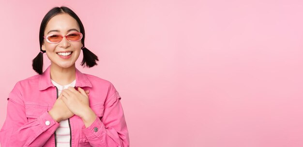 Souriante jeune femme asiatique à lunettes de soleil regardant reconnaissante tenant la main sur le cœur flatté apprécier