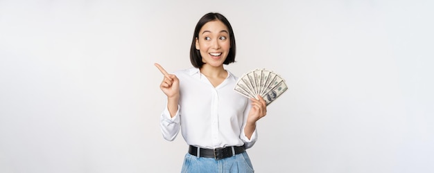 Souriante jeune femme asiatique moderne pointant sur une bannière publicitaire détenant de l'argent comptant en dollars standin