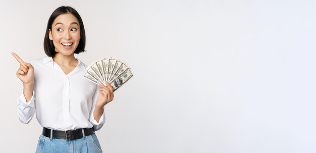 Souriante jeune femme asiatique moderne pointant sur une bannière publicitaire détenant des dollars en espèces debout sur fond blanc