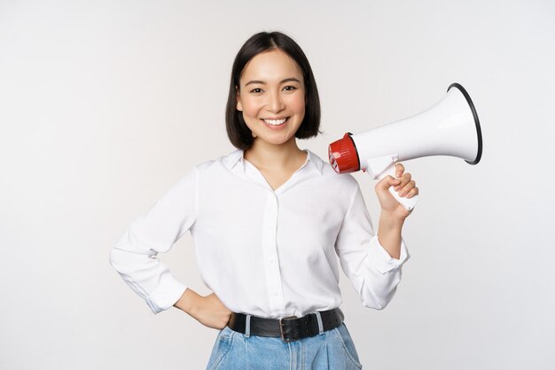 Souriante jeune femme asiatique posant avec le concept de mégaphone d'annonce de nouvelles et d'informations debout sur fond blanc