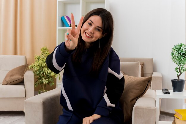 Souriante jeune femme assez caucasienne assise sur un fauteuil dans un salon conçu faisant signe de paix