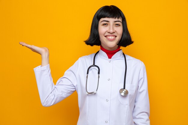 Souriante jeune femme assez caucasienne en uniforme de médecin avec stéthoscope gardant la main ouverte et regardant