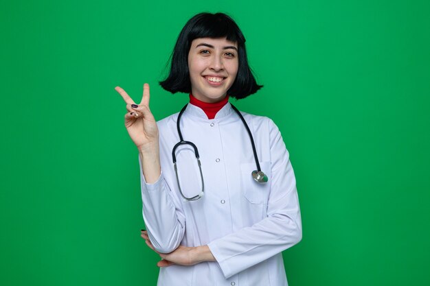 Souriante jeune femme assez caucasienne en uniforme de médecin avec stéthoscope gesticulant signe de victoire