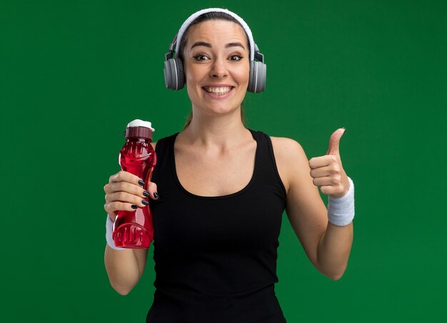 Souriante jeune femme assez sportive portant un bandeau et des bracelets avec un casque tenant une bouteille d'eau regardant à l'avant montrant le pouce vers le haut isolé sur un mur vert