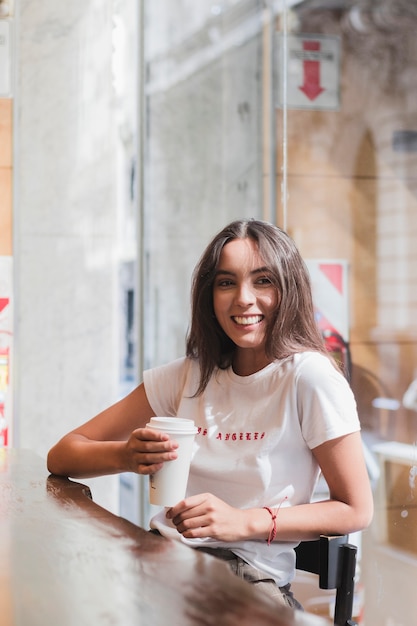 Photo gratuite souriante jeune femme assise dans le café tenant une tasse de café jetable à la main