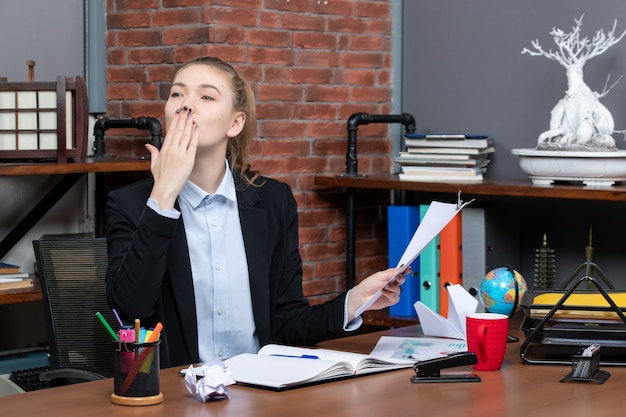 Souriante jeune femme assise à une table et tenant le document envoyant un baiser au bureau