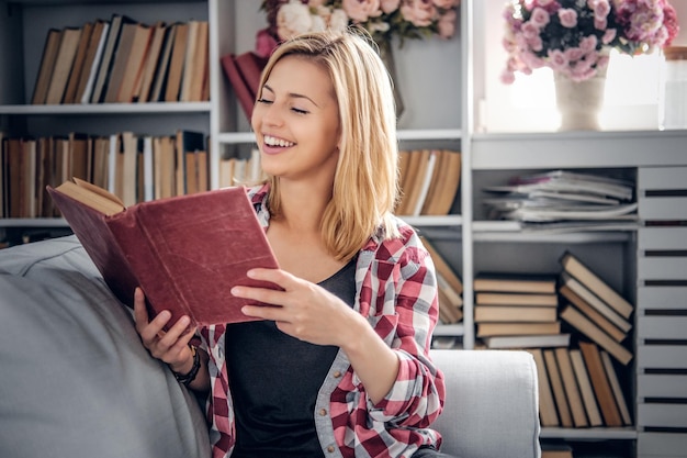 Souriante jeune femme blonde en tenue décontractée tenant un livre, se reposant sur un coach dans un salon.