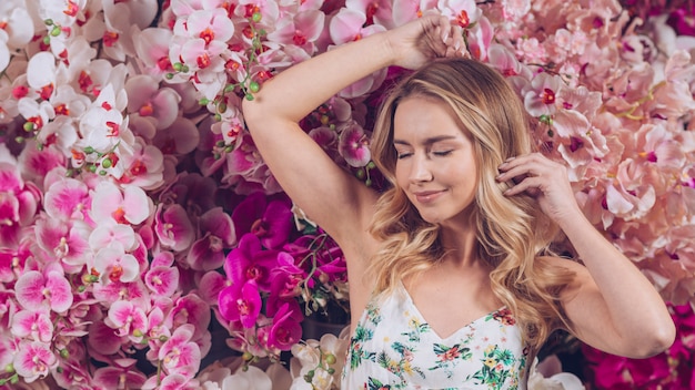 Souriante jeune femme blonde avec des yeux fermés debout contre des orchidées colorées