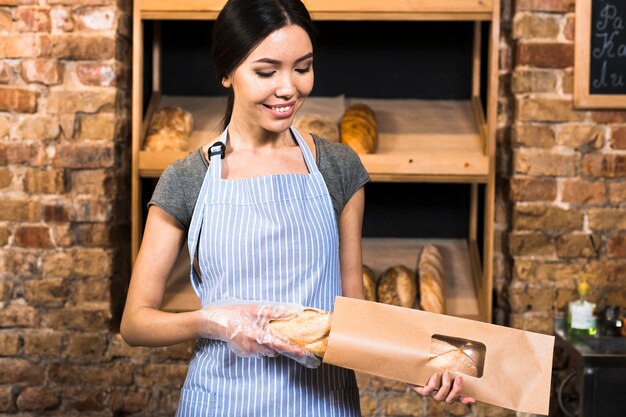 Souriante jeune femme boulangère emballant le pain baguette dans le sac en papier brun du magasin