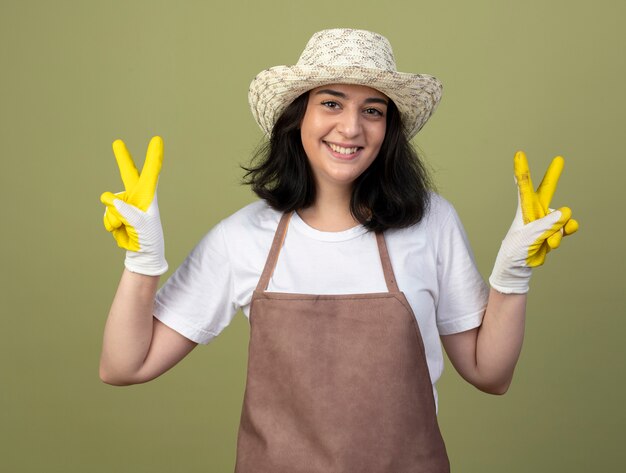 Souriante jeune femme brune jardinière en uniforme portant chapeau de jardinage et gants gestes signe de la main de la victoire avec deux mains isolé sur mur vert olive