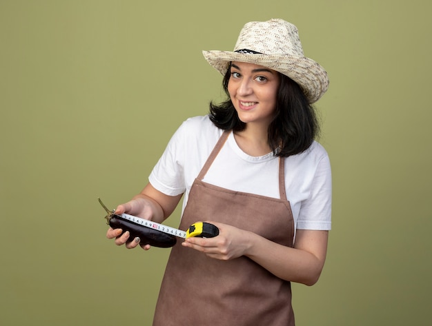 Souriante Jeune Femme Brune Jardinière En Uniforme Portant Chapeau De Jardinage Mesurant L'aubergine Avec Un Ruban à Mesurer Isolé Sur Mur Vert Olive