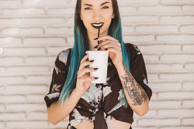 Souriante jeune femme buvant du café avec de la paille contre le mur