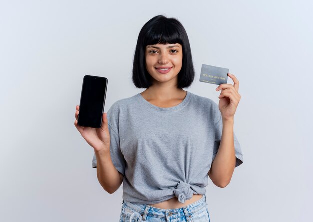 Souriante jeune femme caucasienne brune détient un téléphone et une carte de crédit