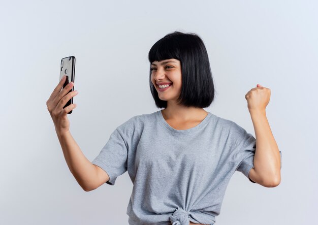 Souriante jeune femme caucasienne brune garde le poing et regarde le téléphone