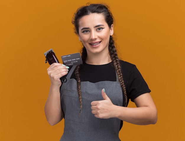 Souriante jeune femme coiffeur en uniforme tenant une carte de crédit et une tondeuse à cheveux montrant le pouce vers le haut isolé sur un mur orange