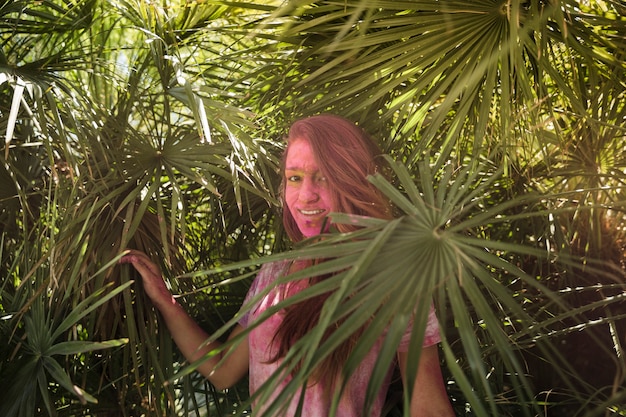 Souriante jeune femme avec la couverture de son visage en couleur holi debout près des feuilles de palmier