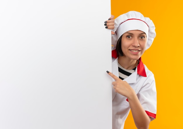 Photo gratuite souriante jeune femme cuisinier en uniforme de chef debout derrière un mur blanc et pointant vers elle isolé sur mur orange