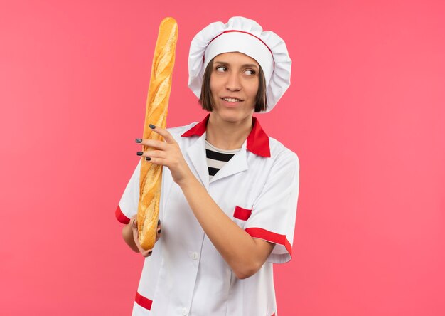 Souriante jeune femme cuisinier en uniforme de chef tenant un bâton de pain à côté isolé sur mur rose