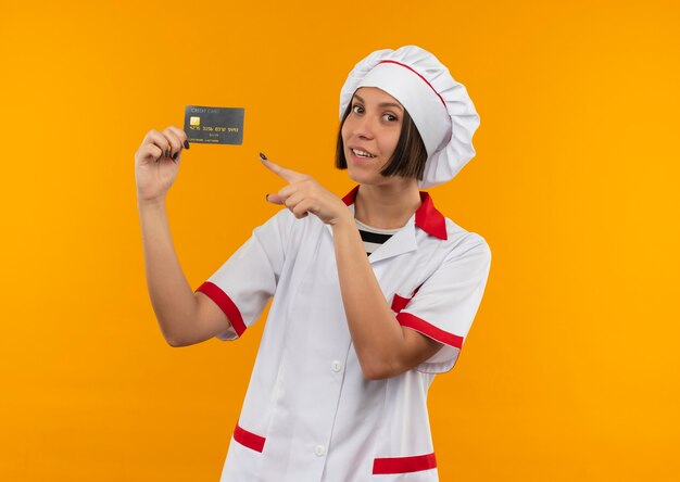 Souriante jeune femme cuisinier en uniforme de chef tenant et pointant sur la carte de crédit à l'avant isolé sur mur orange