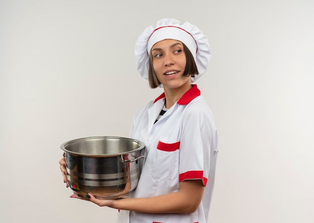 Souriante jeune femme cuisinier en uniforme de chef tenant le pot et regardant à côté isolé sur mur blanc