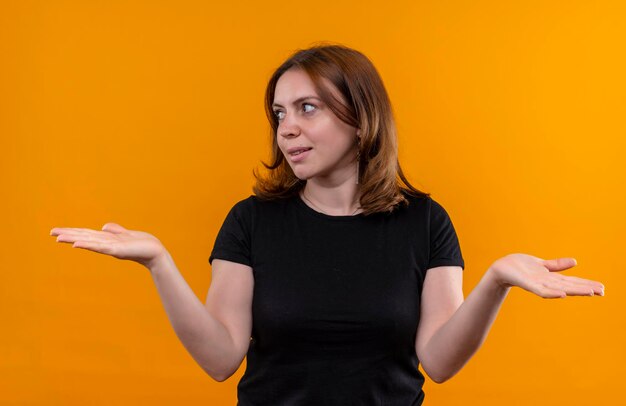 Souriante jeune femme décontractée regardant le côté gauche et montrant les mains vides sur l'espace orange isolé