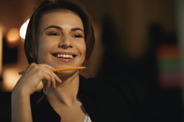 Souriante jeune femme designer assis à l'intérieur la nuit tenant un crayon.