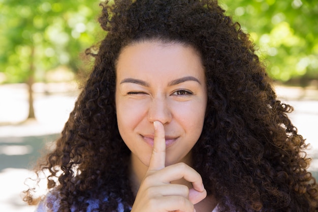 Photo gratuite souriante jeune femme faisant un clin d'oeil et faisant un geste de silence dans le parc