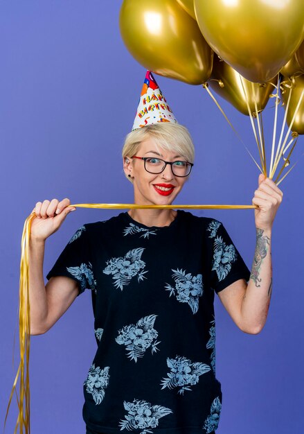 Souriante jeune femme de fête blonde portant des lunettes et une casquette d'anniversaire tenant des ballons à l'avant isolé sur mur violet