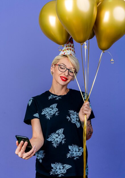 Souriante jeune femme de fête blonde portant des lunettes et une casquette d'anniversaire tenant des ballons et un téléphone mobile prenant selfie isolé sur mur violet