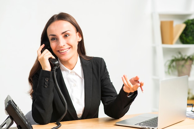 Souriante jeune femme gesticulant tout en parlant au téléphone avec ordinateur portable sur le bureau