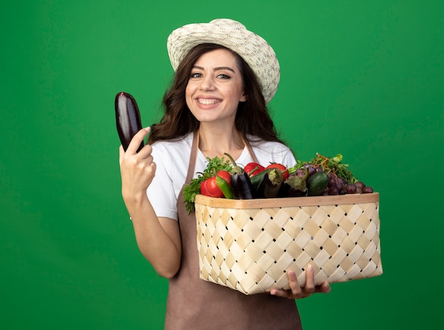 Souriante jeune femme jardinière en uniforme portant chapeau de jardinage détient panier de légumes et aubergines isolé sur mur vert