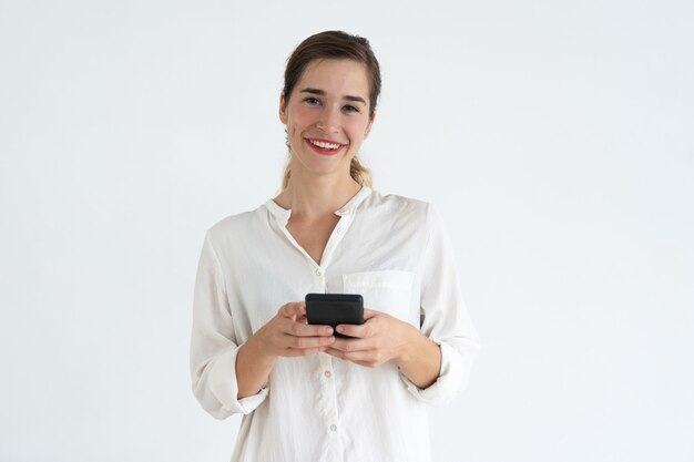 Souriante jeune femme jolie textos sur smartphone et en regardant la caméra.