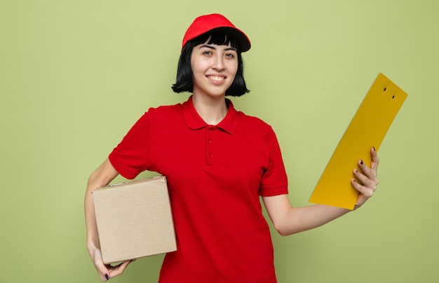 Souriante jeune femme de livraison caucasienne tenant une boîte en carton et un presse-papiers