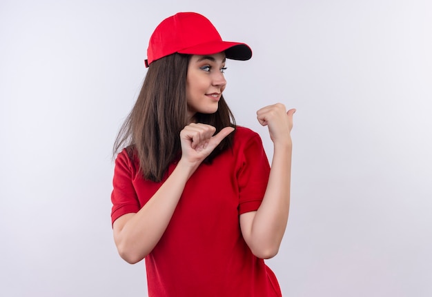 Souriante jeune femme de livraison portant un t-shirt rouge en bonnet rouge pointe vers l'arrière sur un mur blanc isolé