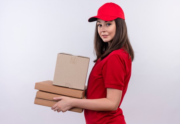 Souriante jeune femme de livraison portant un t-shirt rouge en bonnet rouge tenant une boîte et une boîte à pizza sur un mur blanc isolé