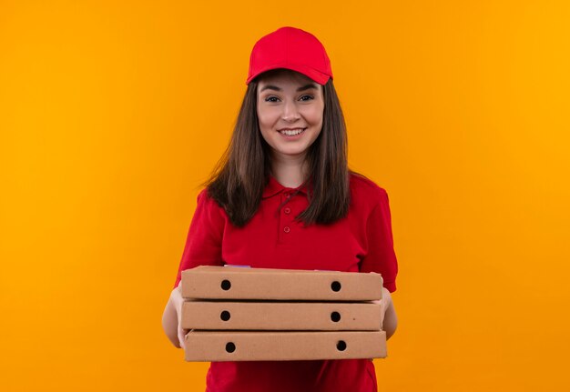 Souriante jeune femme de livraison portant un t-shirt rouge en bonnet rouge tenant une boîte à pizza sur un mur orange isolé