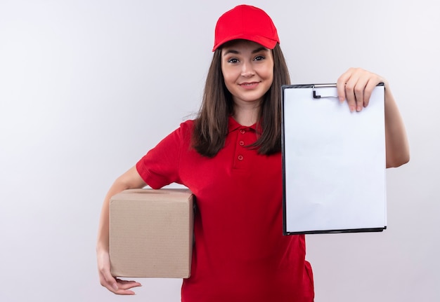 Souriante jeune femme de livraison portant un t-shirt rouge en bonnet rouge tenant une boîte et un presse-papiers sur un mur blanc isolé