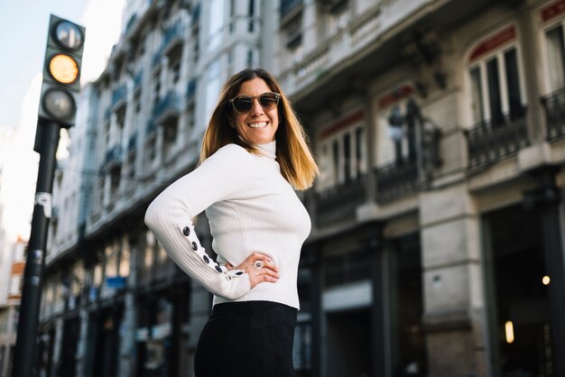 Souriante jeune femme avec des lunettes de soleil près des bâtiments de la ville