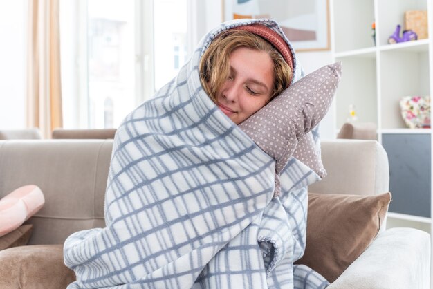 Souriante jeune femme malsaine au chapeau chaud enveloppé dans une couverture souffrant de grippe et de froid tenant un oreiller souriant les yeux fermés assis sur un canapé dans un salon lumineux