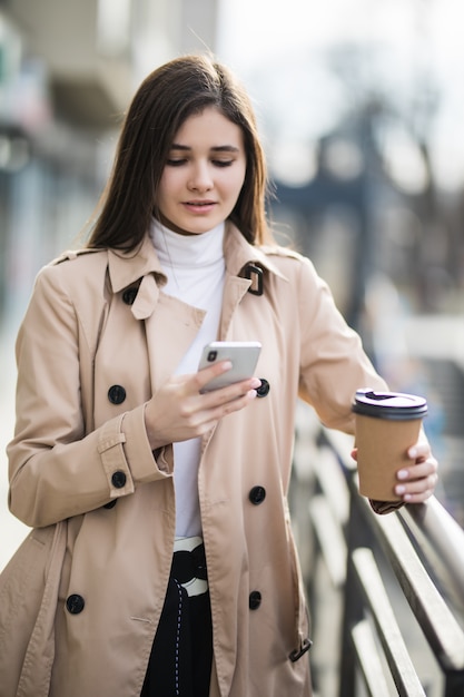 Souriante jeune femme en manteau marron clair lire les nouvelles sur le téléphone à l'extérieur