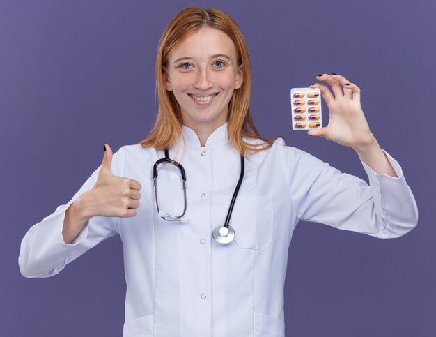 Souriante jeune femme médecin au gingembre portant une robe médicale et un stéthoscope regardant à l'avant montrant un paquet de pilules médicales à l'avant et montrant le pouce vers le haut isolé sur un mur violet