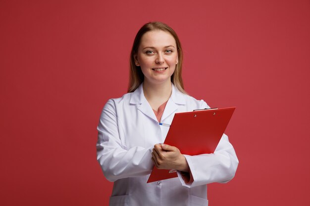 Souriante jeune femme médecin blonde portant une robe médicale et un stéthoscope autour du cou écrit sur le presse-papiers avec un stylo