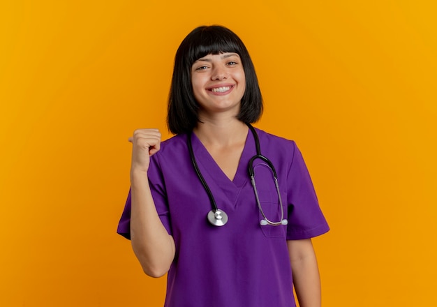 Souriante jeune femme médecin brune en uniforme avec des points de stéthoscope derrière isolé sur fond orange avec copie espace