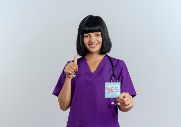 Souriante jeune femme médecin brune en uniforme avec stéthoscope tient oui note et pouces vers le haut