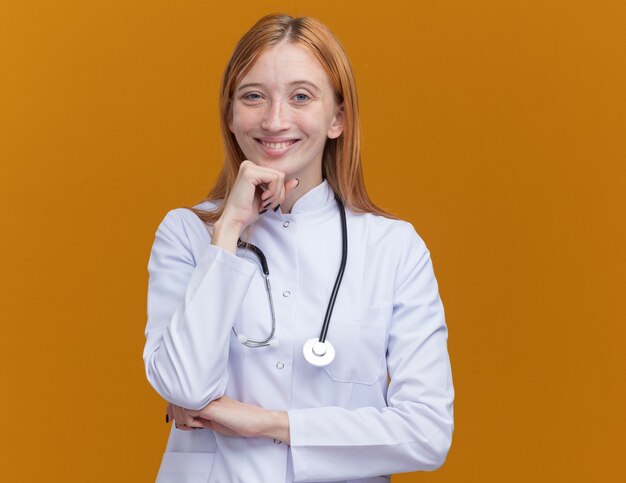 Souriante jeune femme médecin gingembre portant une robe médicale et un stéthoscope gardant la main sur le menton