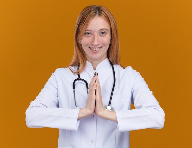 Souriante jeune femme médecin gingembre portant une robe médicale et un stéthoscope gardant les mains ensemble