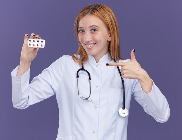 Souriante jeune femme médecin gingembre portant une robe médicale et un stéthoscope regardant l'avant tenant et pointant vers un paquet de pilules médicales isolées sur un mur violet