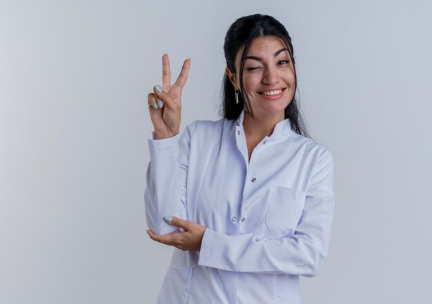 Souriante jeune femme médecin portant une robe médicale faisant signe de paix mettant la main sur le coude isolé sur un mur blanc avec espace de copie